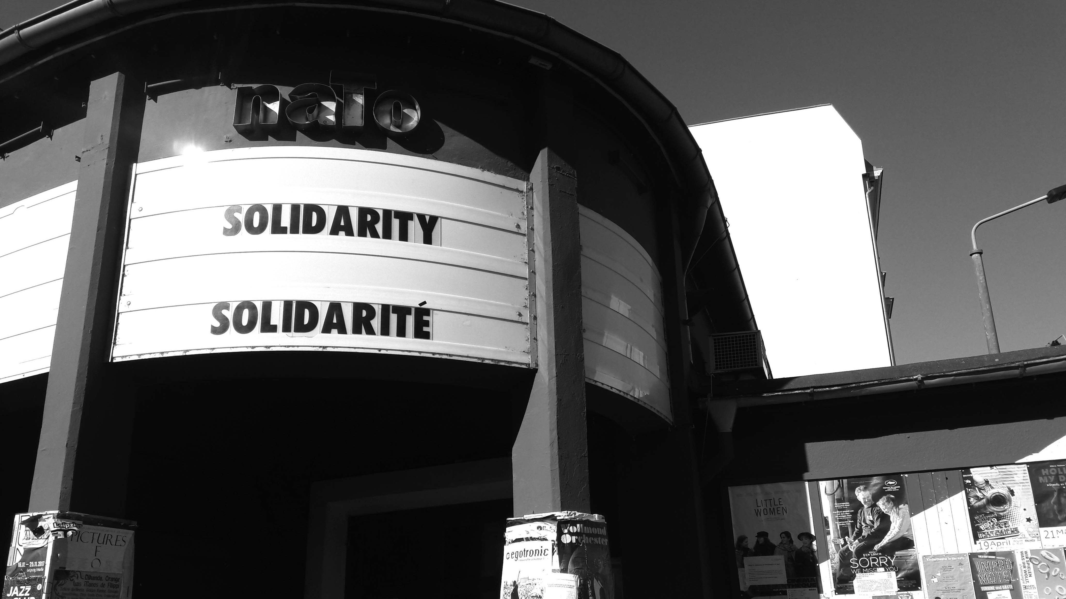 Schwarz-Weiß-Aufnahme der Spielstätte naTo von außen, an der Anzeige über dem Eingang ist der Aufruf "Solidarity / Solidarité" angeschlagen